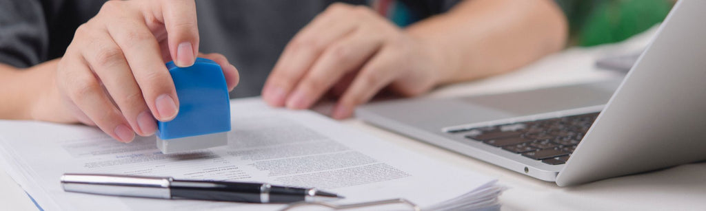 A person at a desk with papers, a pen and a laptop. He is stamping a document.