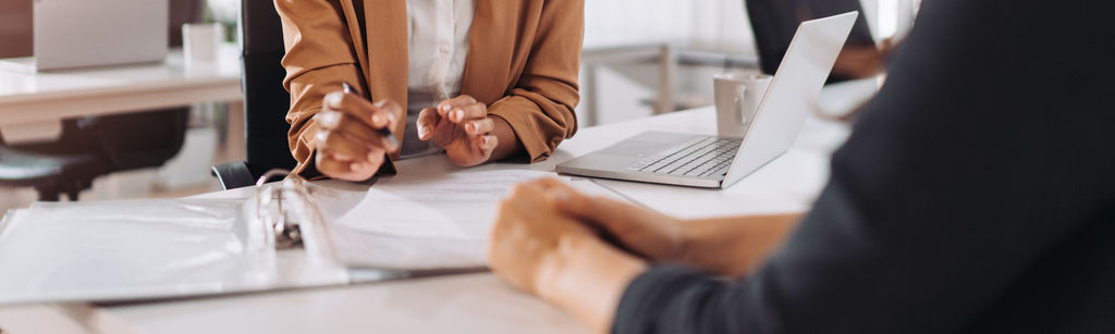 Two people sitting at a table that has a laptop and various papers placed on it.