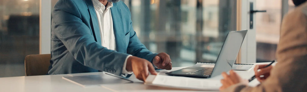 A person handing someone a document to someone. There is a laptop on the table.